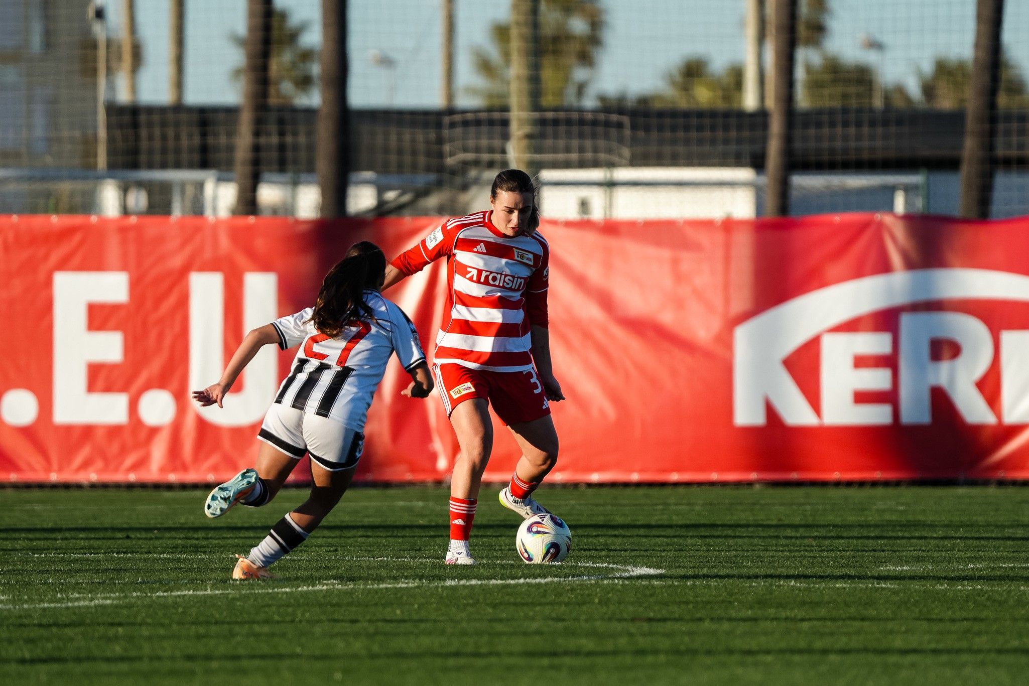 Zwei Fußballspielerinnen auf einem Spielfeld, eine in rot-weiß gestreifter Uniform dribbelt den Ball, während die andere sie verfolgt.