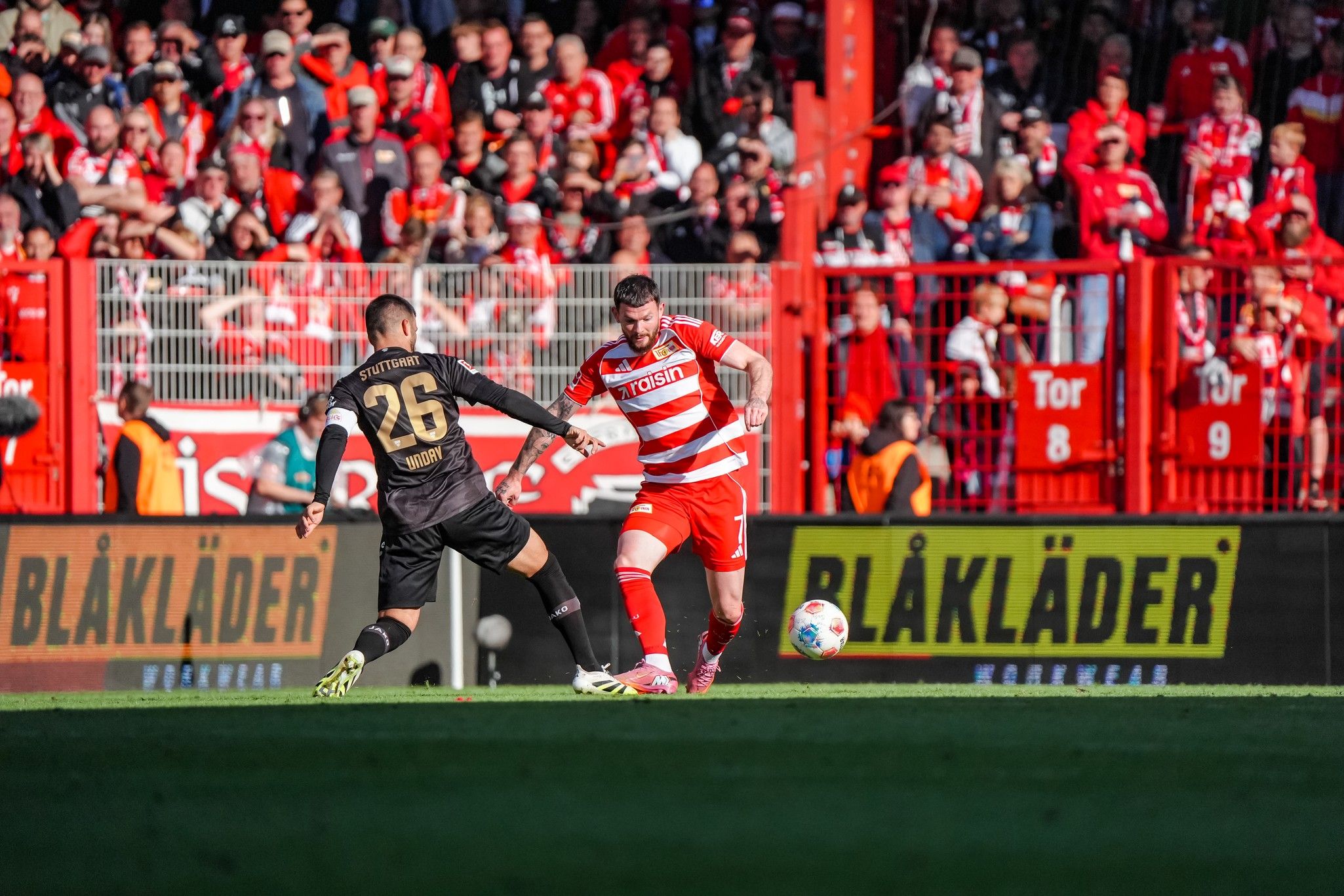 Fußballspiel mit zwei Spielern im Mittelpunkt, der eine in gestreiften roten und weißen Trikots, der andere in schwarzer Ausrüstung.
