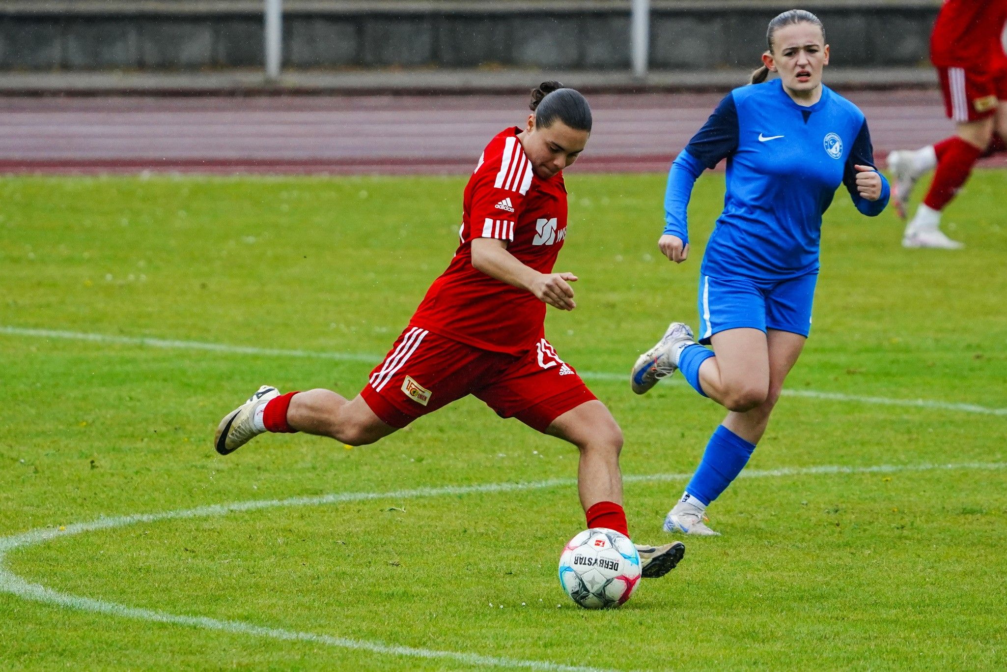 Spielende Frauen im Fußball: Eine Spielerin in roter Uniform kickt den Ball, während eine Gegnerin in Blau hinterherläuft.