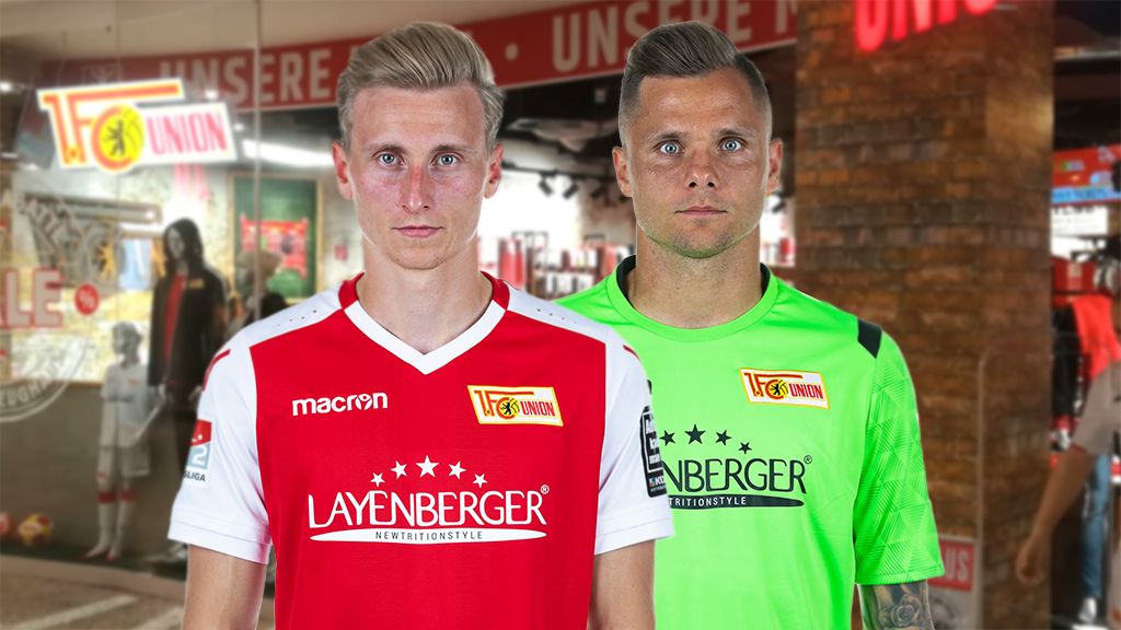 Zwei Fußballspieler in Trikots des 1. FC Union Berlin, einer in rotem und einer in grünem Outfit, stehen vor einem sportlichen Hintergrund.