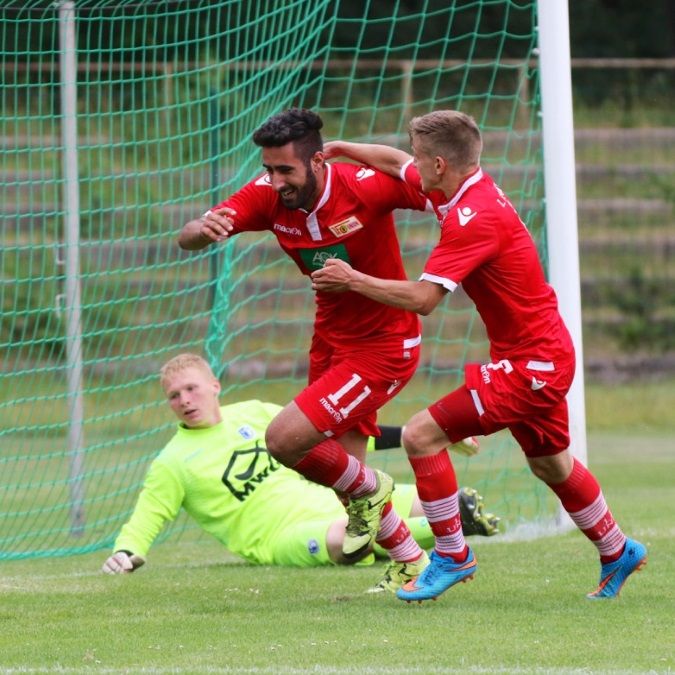 Zwei Fußballspieler in roten Trikots feiern einen Torerfolg, während ein Torwart im Hintergrund enttäuscht aussieht.