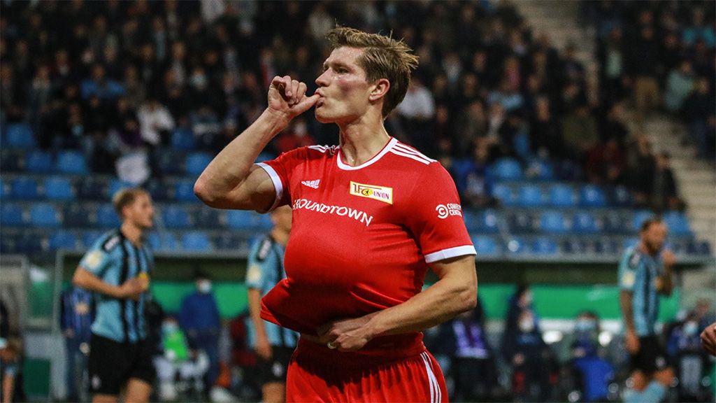 A soccer player in a red jersey makes a referee gesture with his hand while standing on the field.