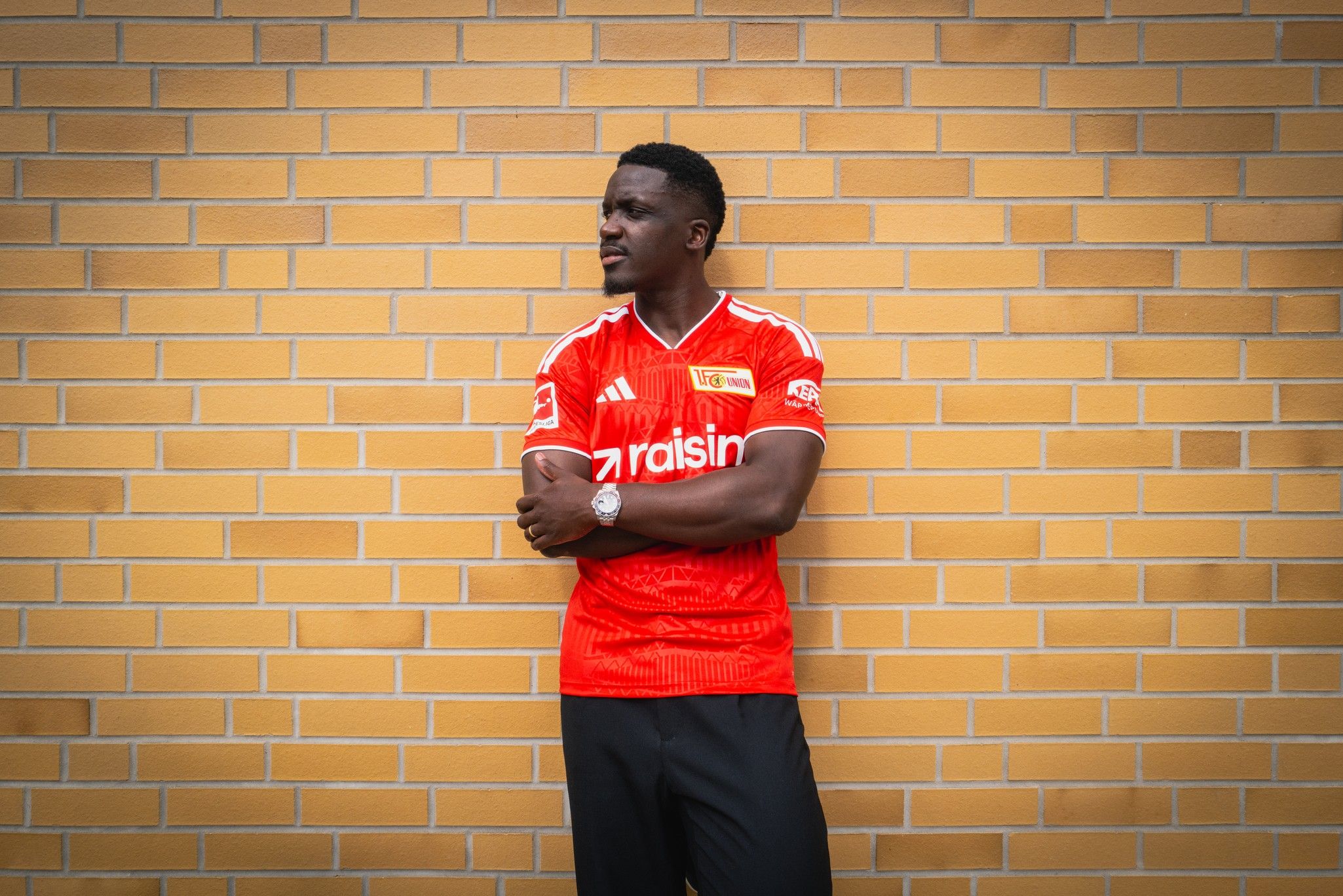 Ein Mann steht mit verschränkten Armen an einer gelben Ziegelwand und trägt ein rotes Fußballtrikot.