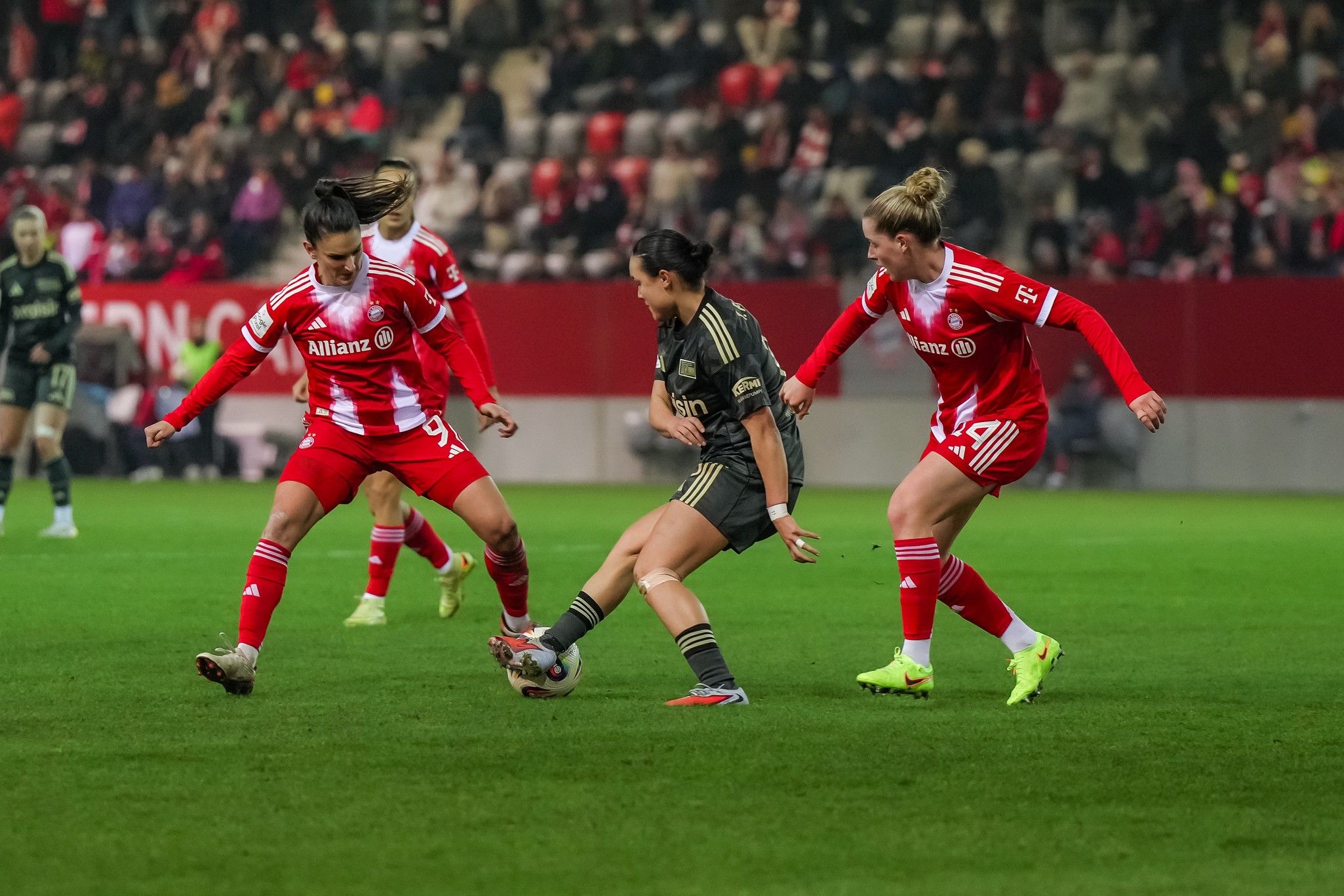 Zwei Spielerinnen in roten Trikots versuchen, eine gegnerische Spielerin in einem schwarzen Trikot am Ball zu hindern.