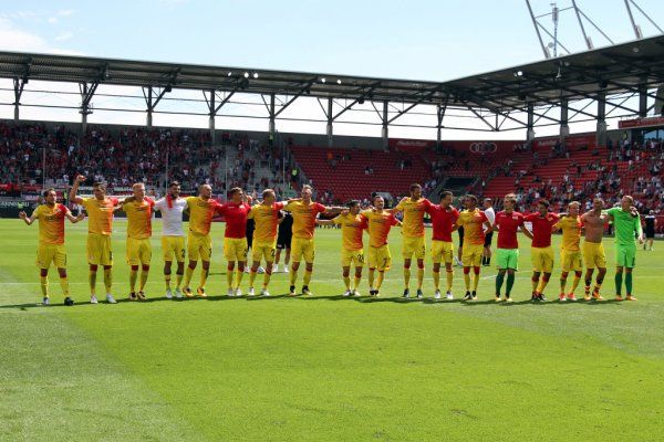 A soccer team is lined up on the field celebrating after a game. Spectators are seen in the background.