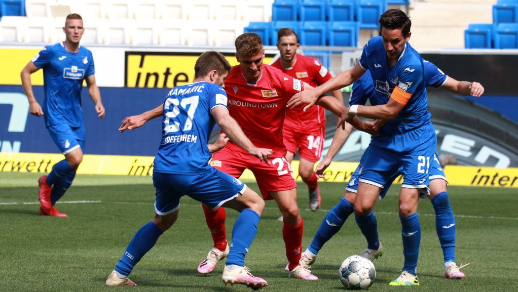 Zwei Spieler in blauen Trikots umkämpfen den Ball gegen einen Spieler in rotem Trikot auf einem Fußballfeld.