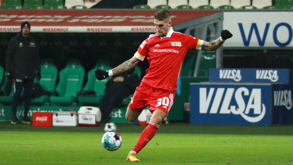 A player of 1. FC Union Berlin in a red jersey kicks the ball during a soccer match on a green field.