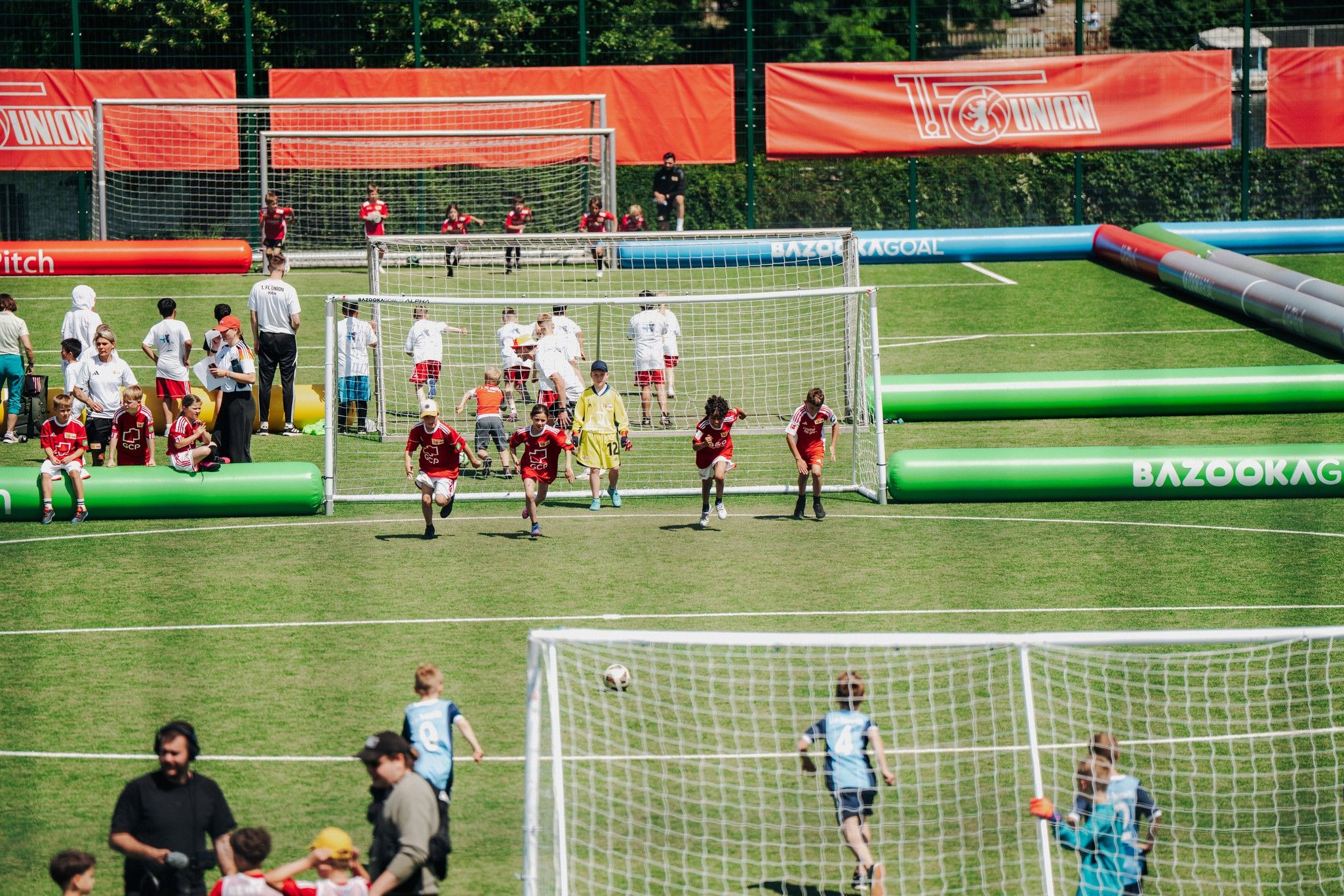 Eine Sportanlage mit Fußballspielern auf dem Feld, Fans auf der Tribüne und Kindern, die spielen. Bunte Bänke im Hintergrund sichtbar.