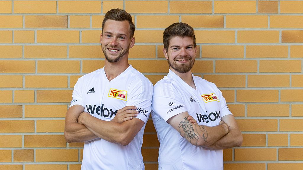 Two soccer players are smiling back to back in white jerseys in front of a yellow brick wall.