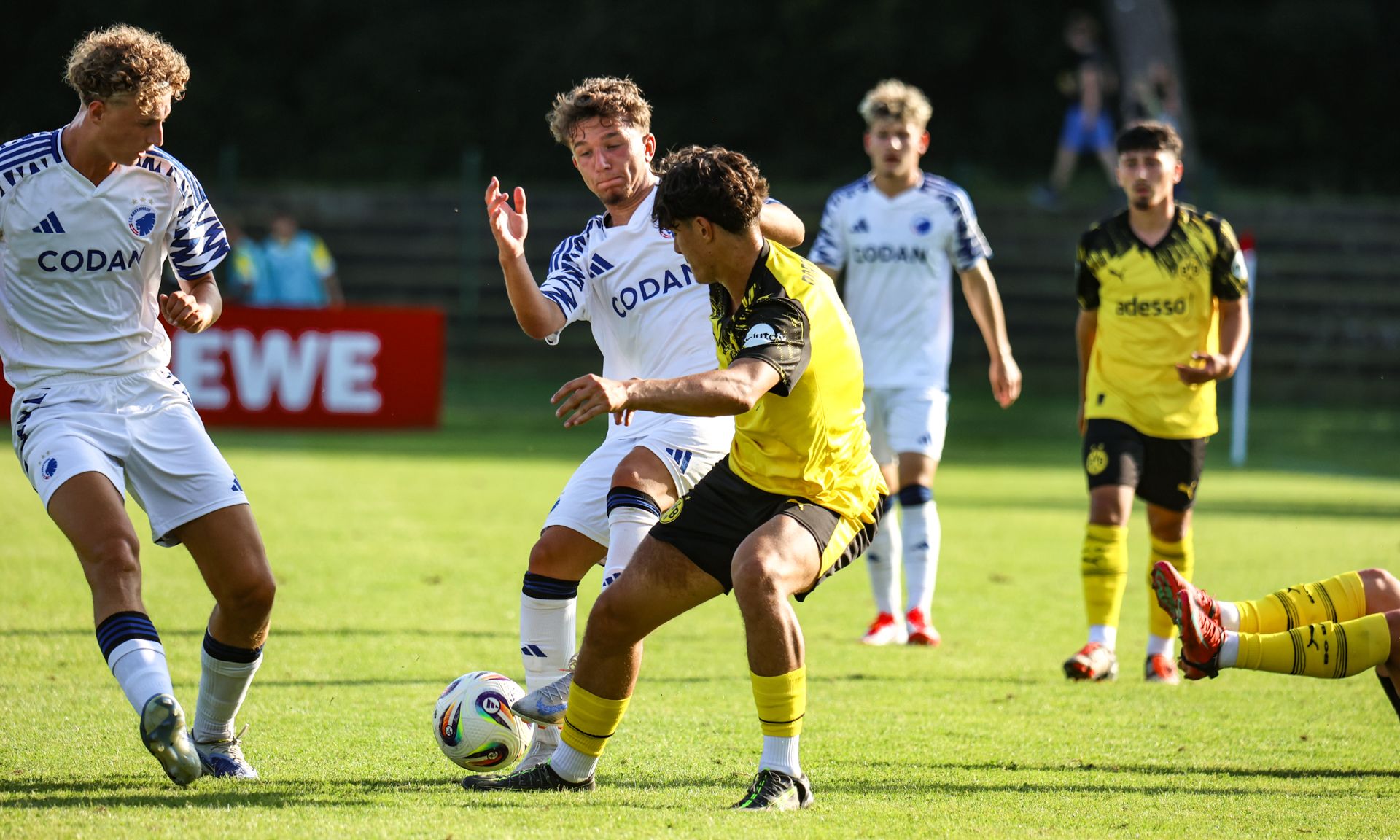 Zwei Fußballspieler kämpfen im Spiel um den Ball, während weitere Spieler im Hintergrund beobachten.