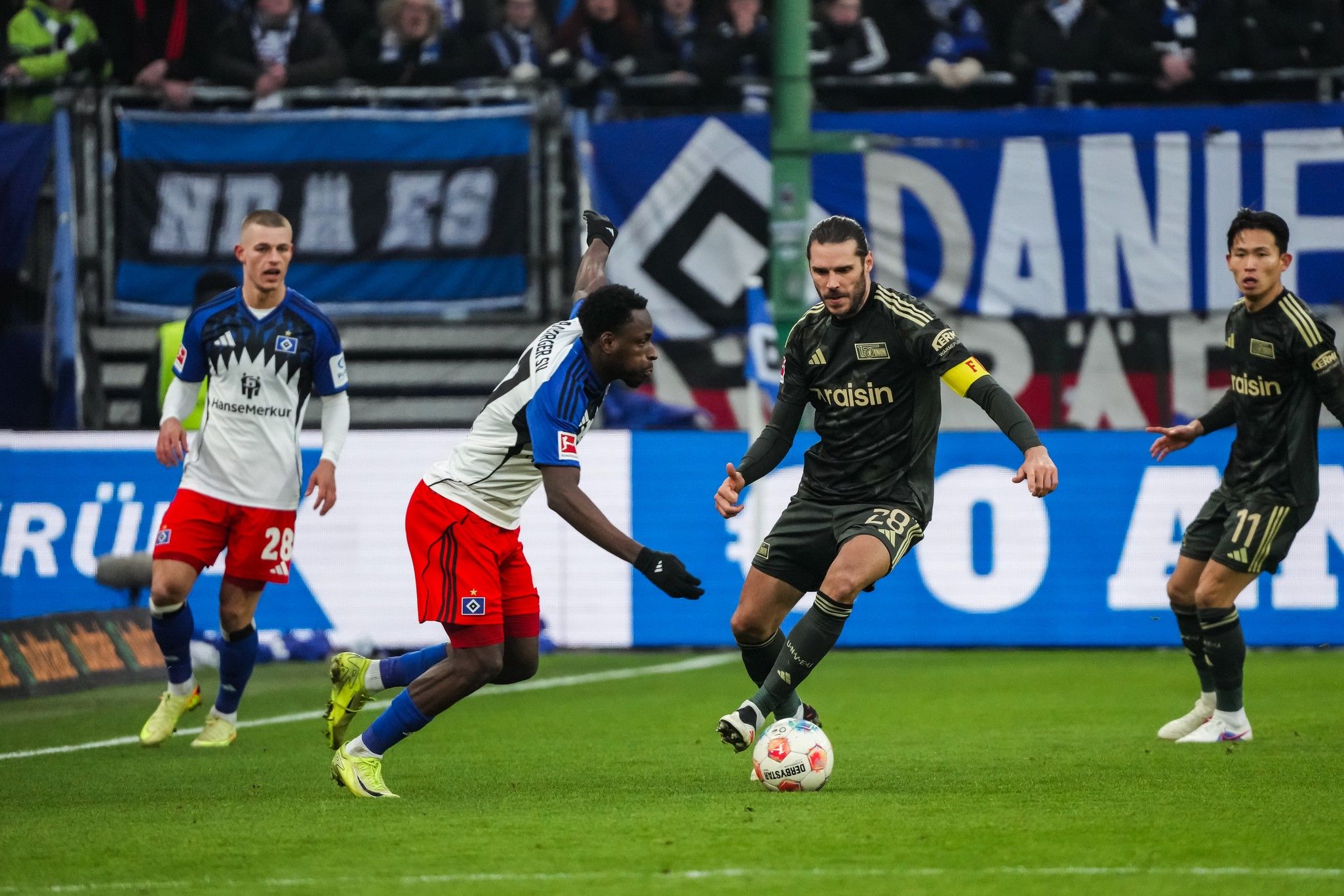 Zwei Fußballspieler im Wettkampf um den Ball, Zuschauer im Hintergrund. Ein Spieler trägt ein rotes und weißes Trikot, der andere ein schwarzes.