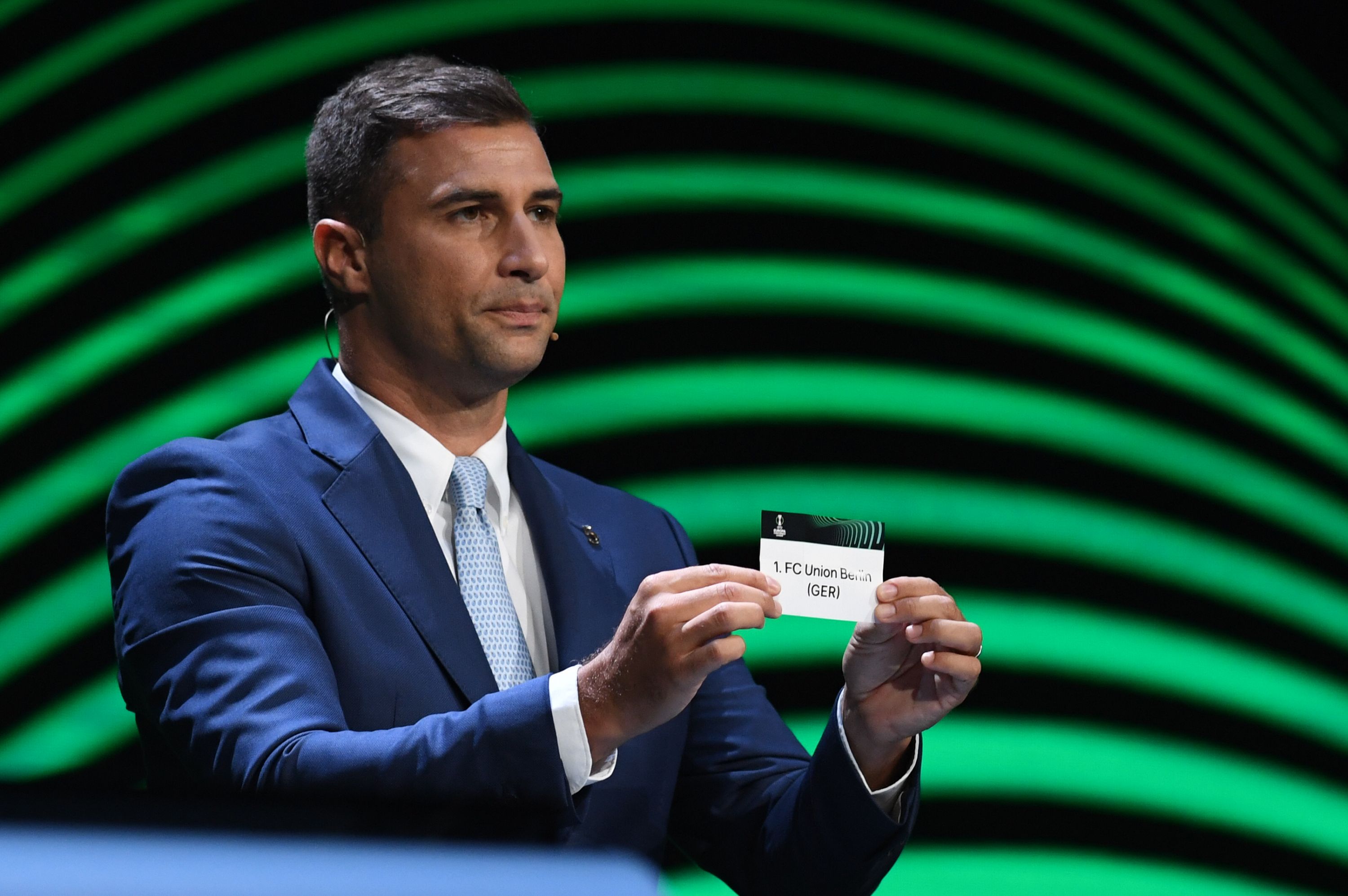 A man in a blue suit holds a note with the name "1. FC Union Berlin" in front of him, with green waves in the background.