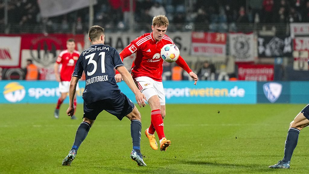 Ein Fußballspieler in rotem Trikot dribbelt den Ball, während ein Gegner in dunkler Kleidung ihn verteidigt.