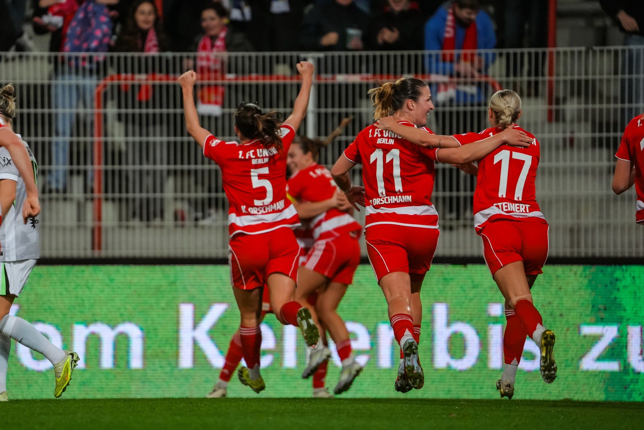Spielerinnen eines Fußballteams in roten Trikots jubeln nach einem Tor, während im Hintergrund Zuschauer sichtbar sind.