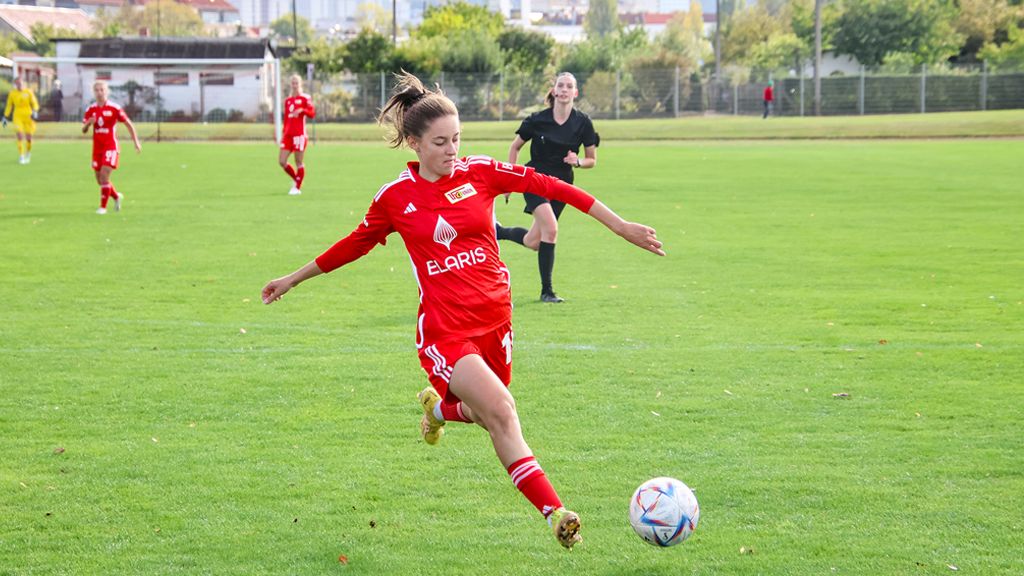A player in a red jersey kicks the ball on a soccer field while other players stand in the background.