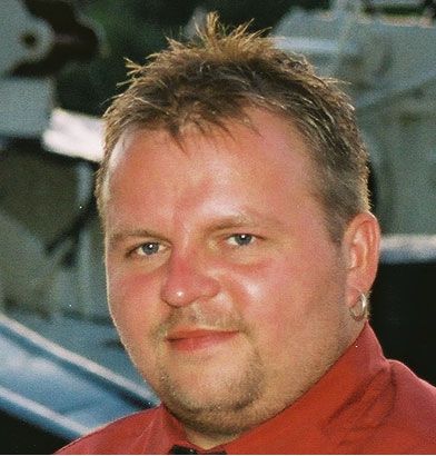 Ein Mann mit kurzen, hellbraunen Haaren und blauen Augen lächelt, trägt ein rotes Hemd und einen Ohrring.