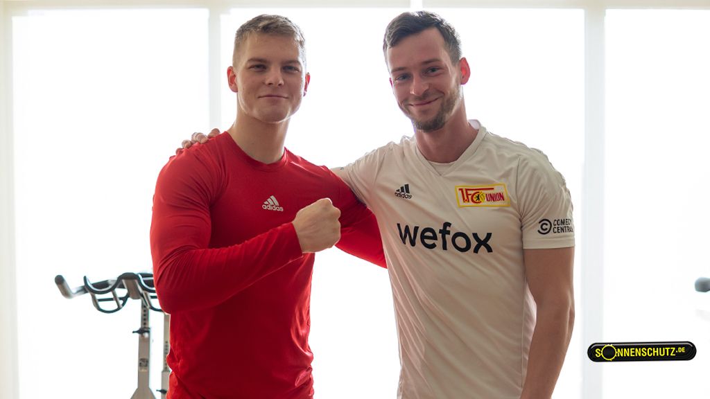 Zwei sportliche Männer stehen lächelnd nebeneinander in einem hellen Raum, einer in rotem Shirt, der andere in einem weißen Trikot.