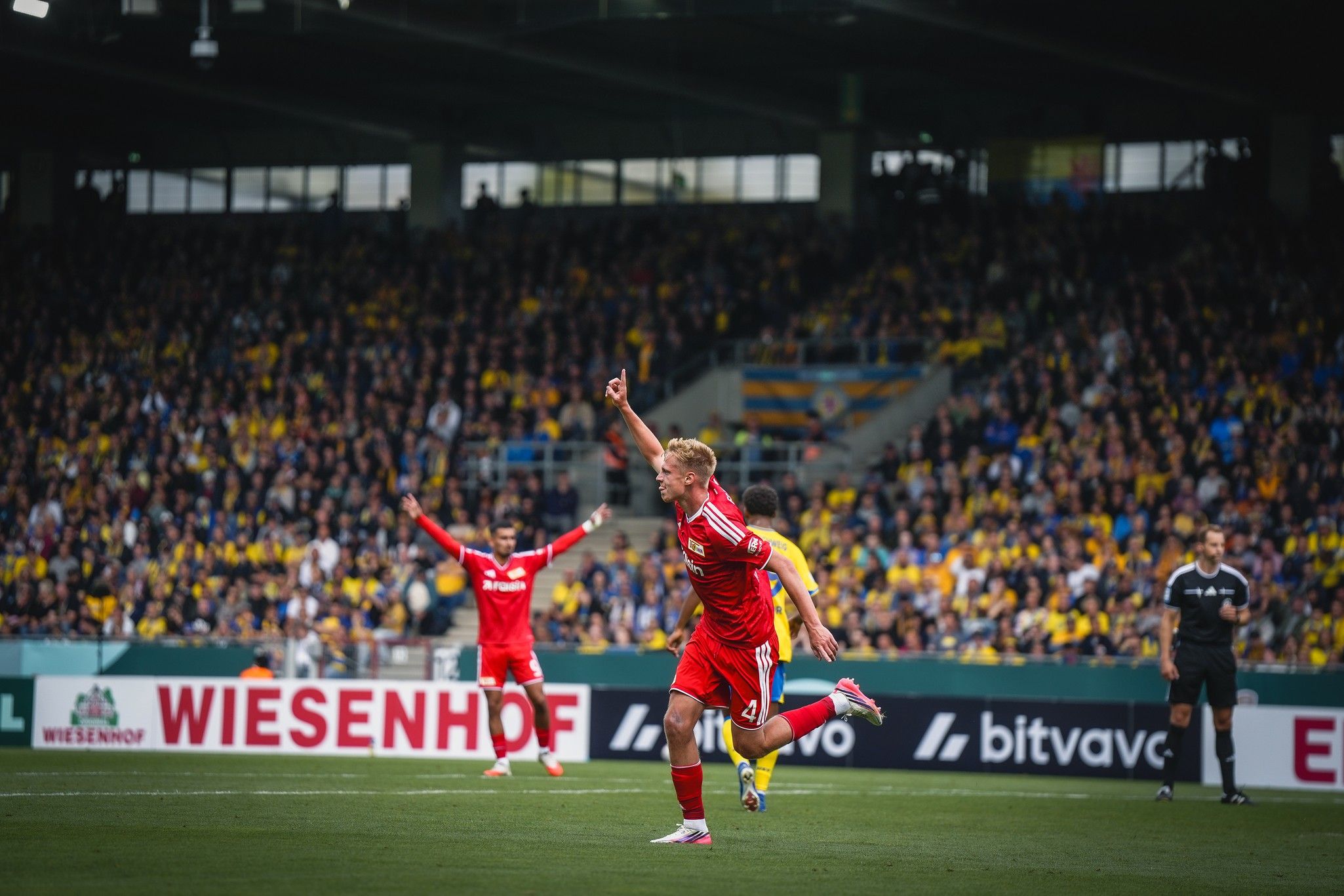Ein Spieler in rotem Trikot jubelt im Stadion, während im Hintergrund ein weiterer Spieler feiert und die Zuschauer applaudieren.