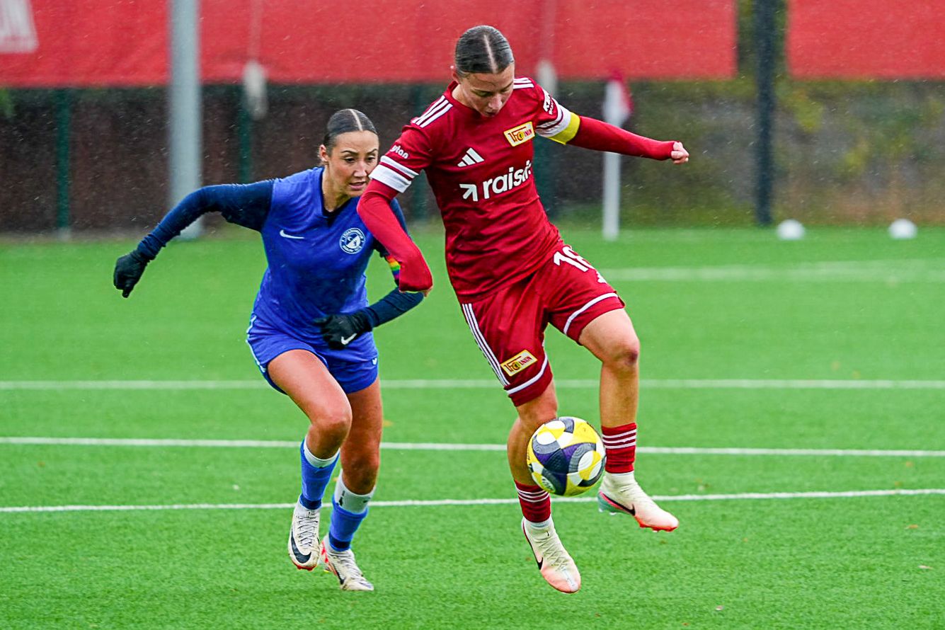 Zwei Fußballspielerinnen im Wettkampf auf einem Rasenfeld, eine in blauer und die andere in roter Uniform, während es regnet.