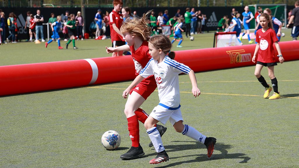 Zwei Mädchen spielen Fußball auf einem Kunstrasenplatz, umgeben von Zuschauern und anderen Kindern.