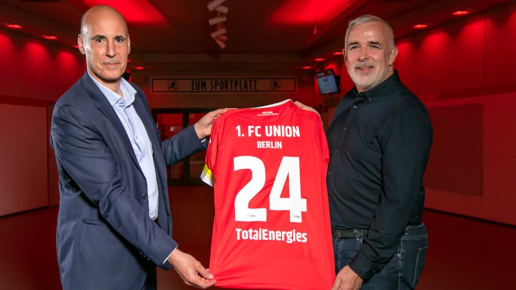 Two men are holding a red-and-white football jersey with the number 24 and the TotalEnergies logo.