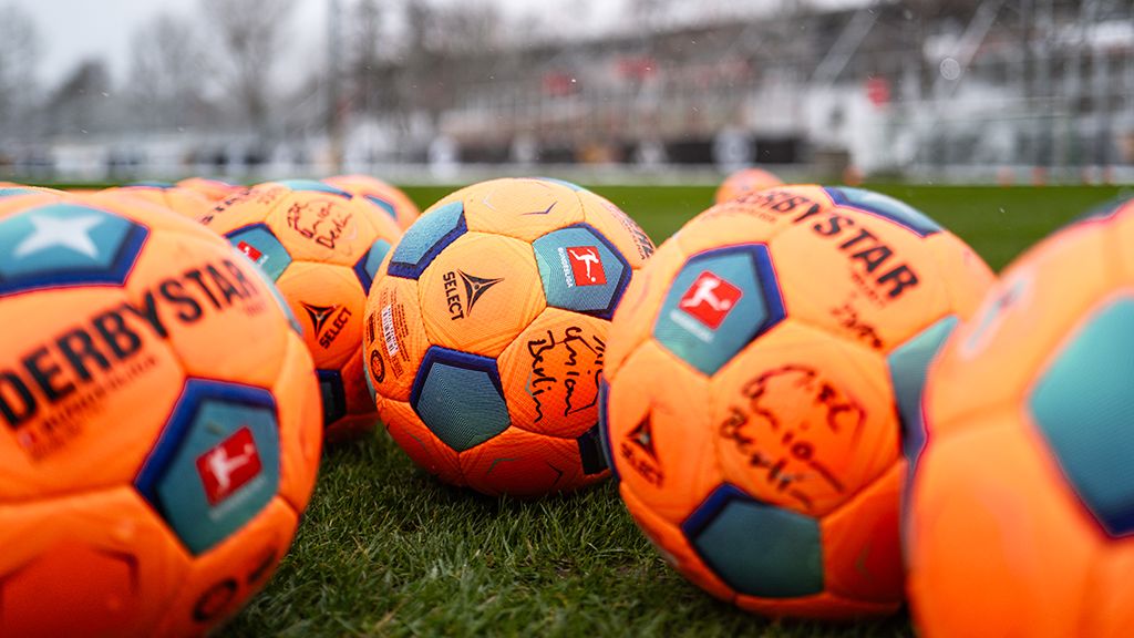 Mehrere orangefarbene Fußball mit blauen und weißen Details liegen auf einem grünen Rasenfeld. Einige Bälle sind signiert.