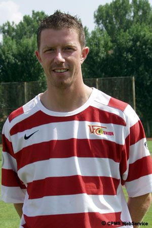 Ein junger Mann in einem rot-weiß gestreiften Fußballtrikot steht im Freien vor einem Sportfeld.