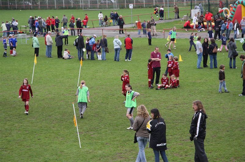 Kinder spielen Fußball auf einem Sportplatz, während Zuschauer und andere Aktivitäten im Hintergrund zu sehen sind.