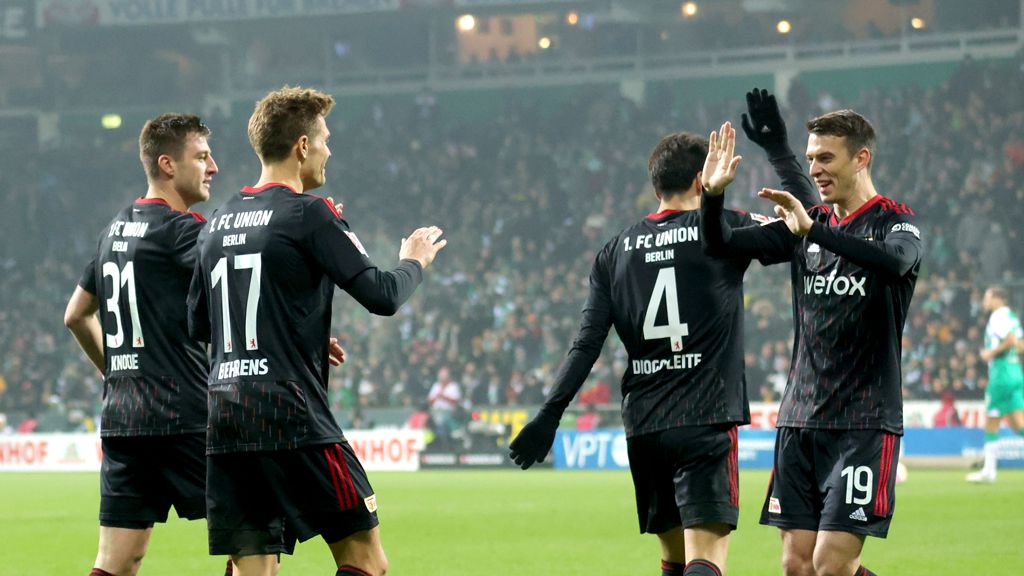 Football team in black jerseys celebrates a goal on the field. Spectators in the background.