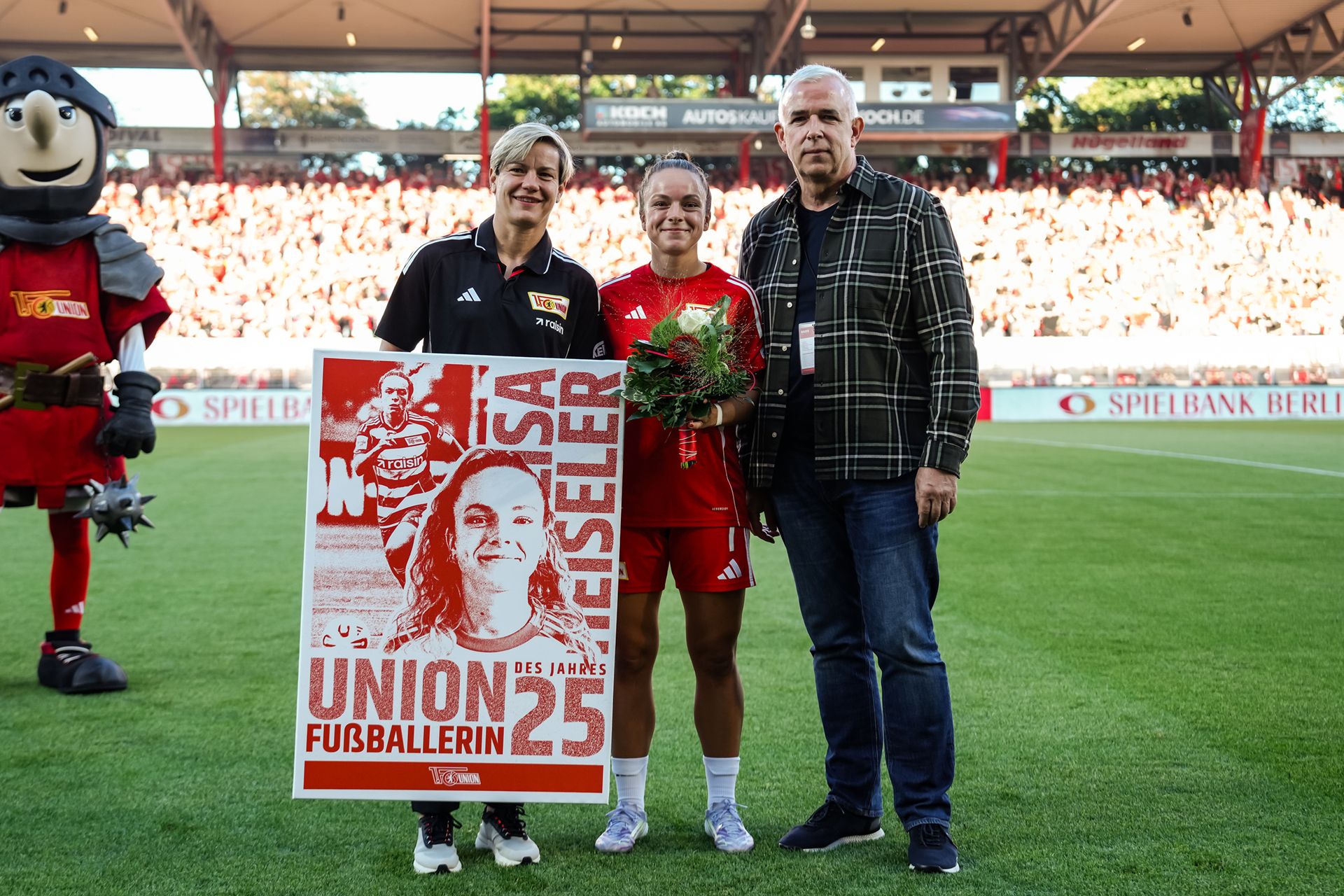 Zwei Personen halten ein Plakat mit einer Spielerin und Blumen, umrahmt von einem Sportmaskottchen im Stadion.