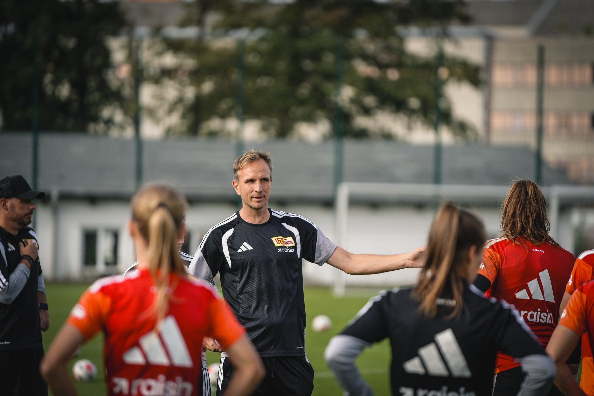 Ein Trainer spricht während eines Fußballtrainings mit Spielerinnen im Vordergrund, die in roten und schwarzen Trikots gekleidet sind.