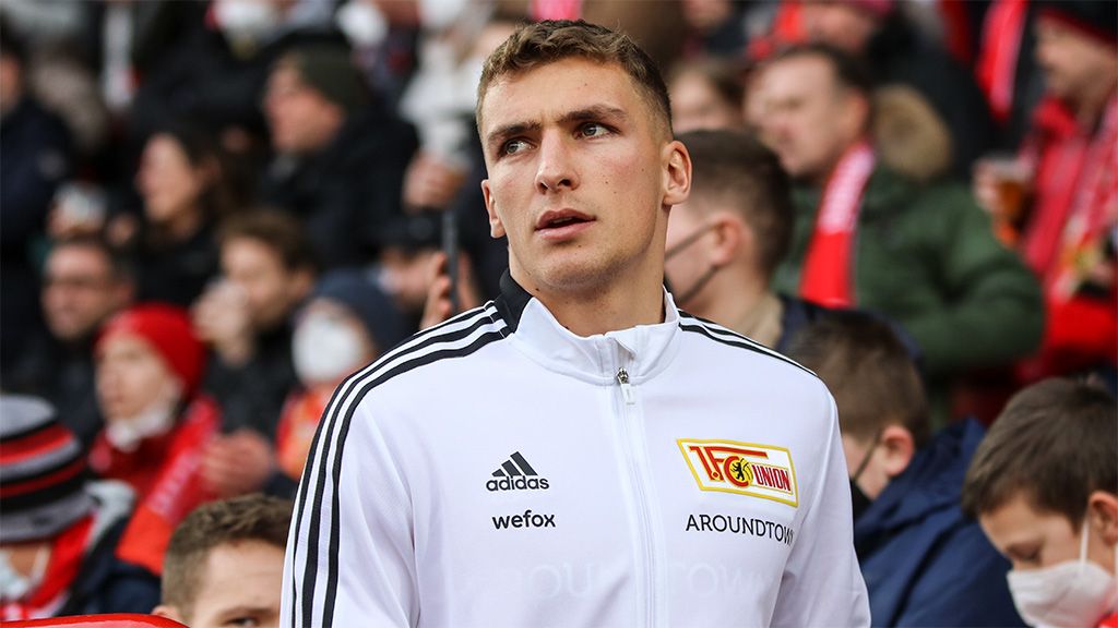 A young football player in a white tracksuit with black details stands in a crowd in the stadium.