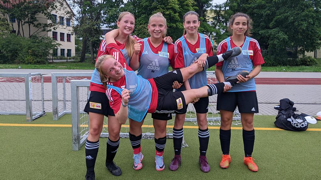 Fünf junge Fußballspielerinnen posieren fröhlich auf dem Platz, einige lachen und eine wird spielerisch gehalten.