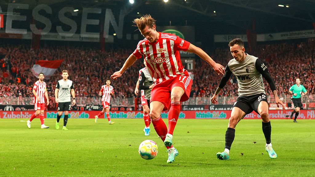 Ein Fußballspieler mit roten und weißen Streifen dribbelt den Ball, während ein Gegner in grauer Uniform versucht, ihn zu stoppen.