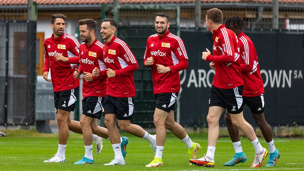 Sechs Fußballspieler in roten Trikots joggen auf einem Trainingsplatz.