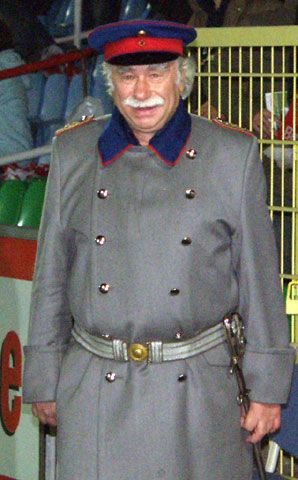 Ein älterer Mann in einem grauen Militärmantel mit blauen Kragen und rotem Hut steht vor einer Tribüne.