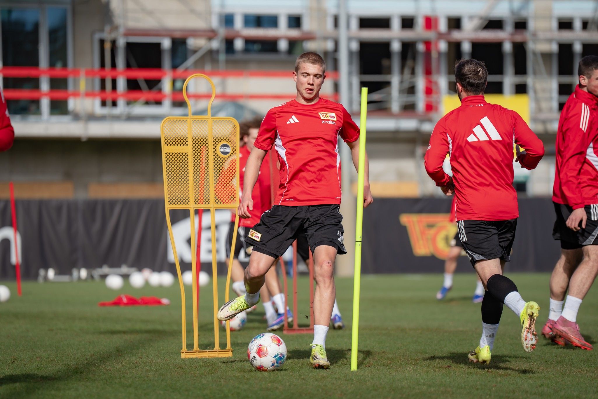 Ein Fußballspieler in rotem Trikot trainiert auf einem Platz, umgeben von Trainingsgeräten und anderen Spielern.