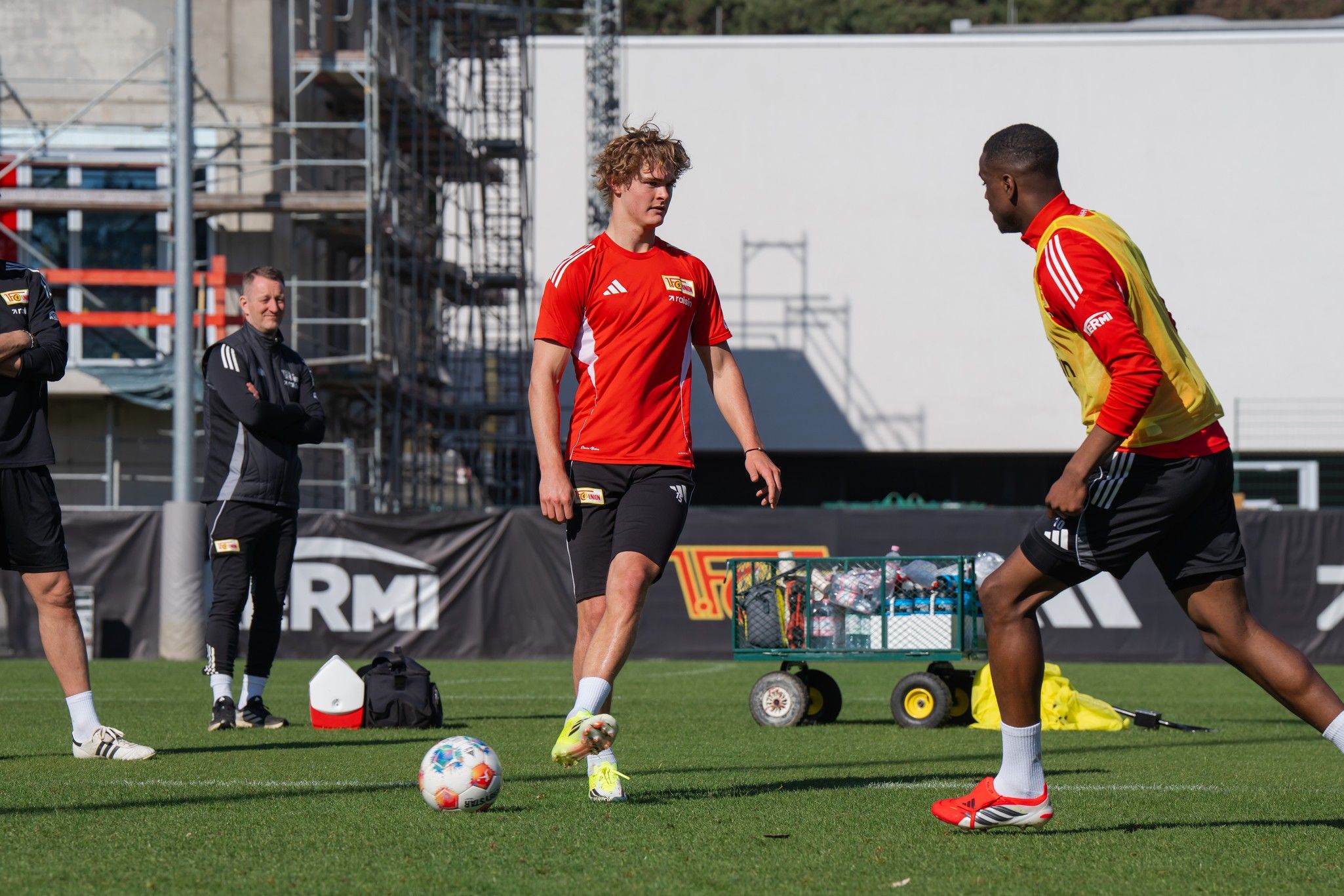 Zwei Fußballspieler in Training, ein Spieler dribbelt den Ball, während ein anderer sich auf den Pass vorbereitet. Im Hintergrund Trainer und Ausrüstung.