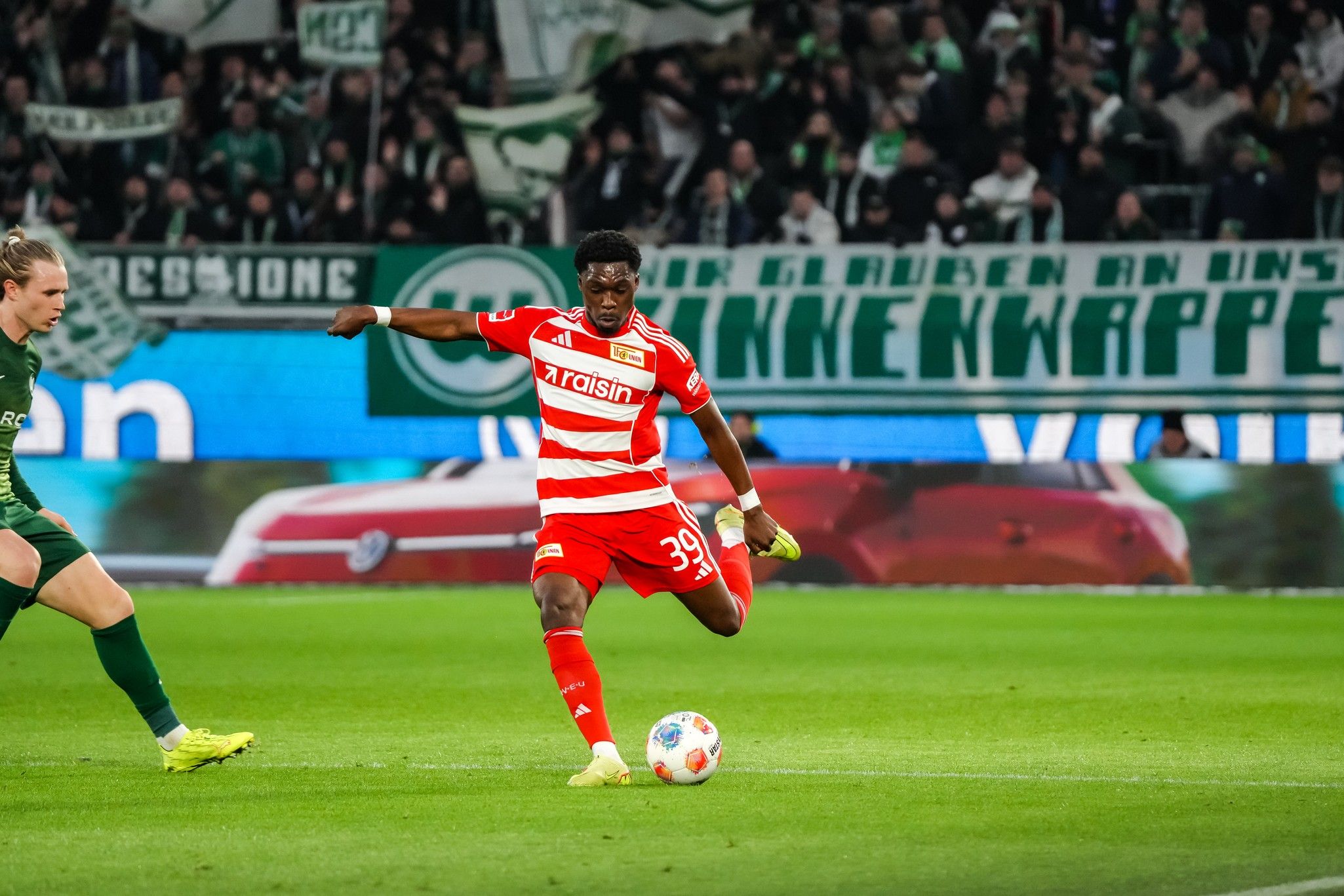 Spielszene im Fußball mit einem Spieler in rot-gestreifter Uniform, der den Ball an einem verdeckten Tor vorbeispielt. Fans im Hintergrund.