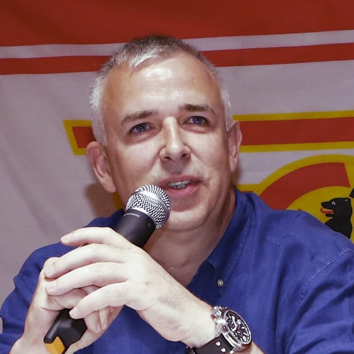 Ein Mann mit grauen Haaren spricht in ein Mikrofon, er trägt ein blaues Hemd und sitzt vor einem Hintergrund mit einer Flagge.