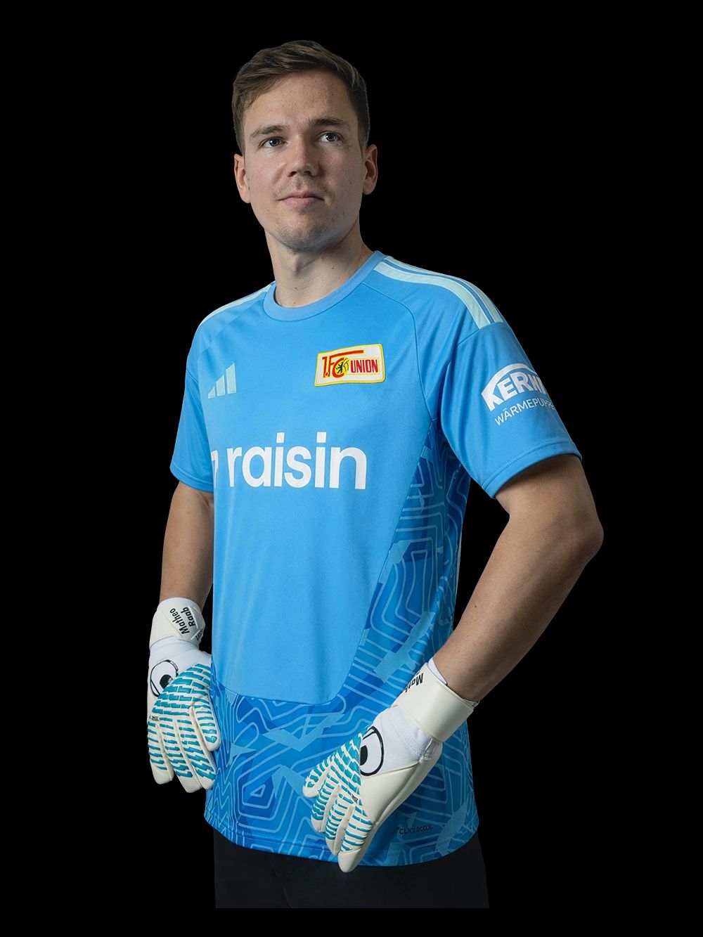 Ein Torwart in einem blauen Trikot mit dem Logo eines Vereins, steht mit verschränkten Armen und trägt Torwarthandschuhe.