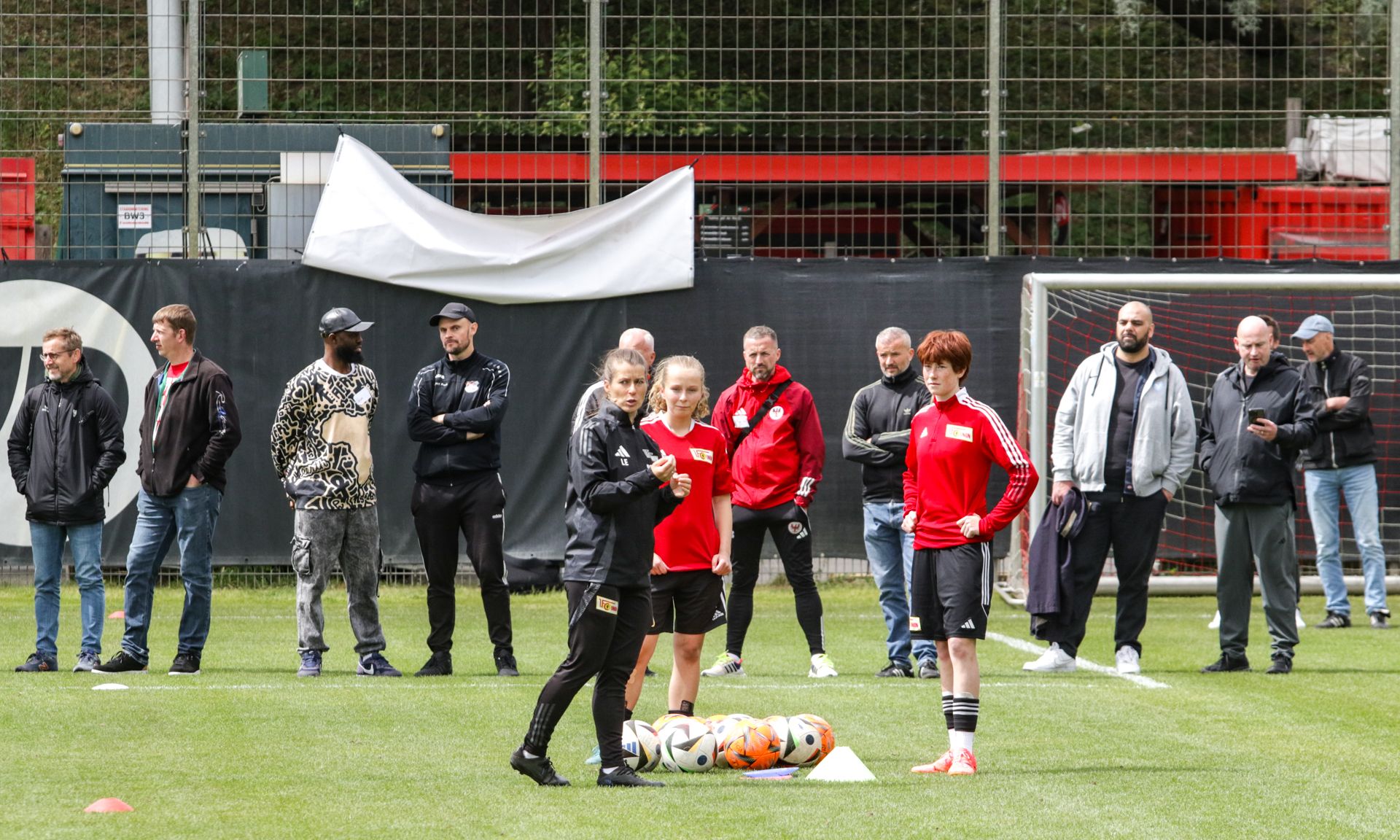 Training auf einem Fußballplatz mit Trainerinnen und Spielerinnen, umgeben von Zuschauern in sportlicher Kleidung.