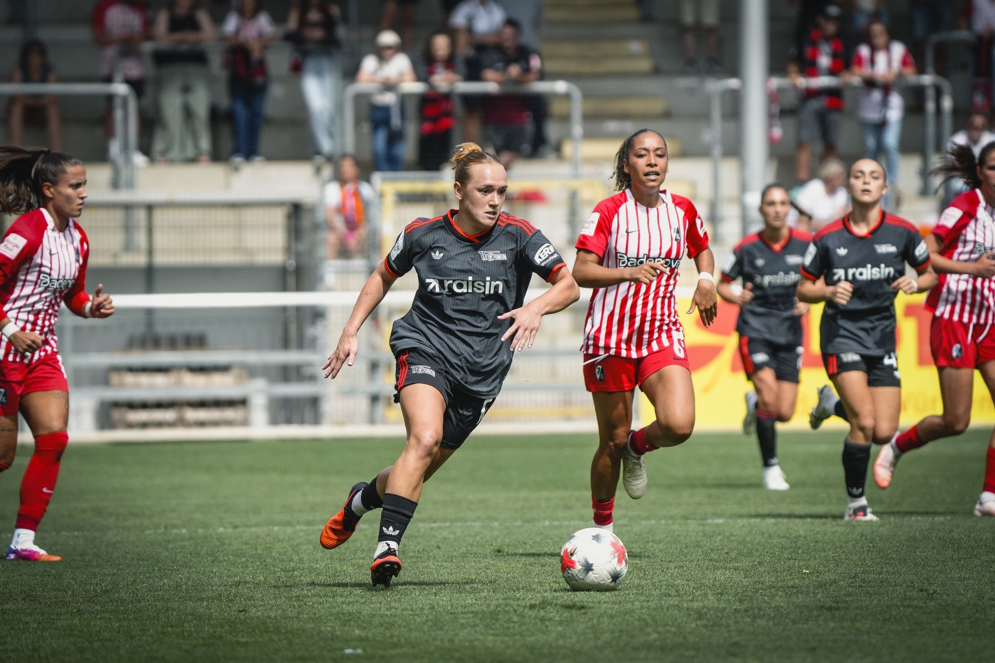 Zwei Frauenfußballspielerinnen rennen in einem Stadion, eine in grauem und eine in rot-weiß gestreiftem Trikot. Zuschauer im Hintergrund.