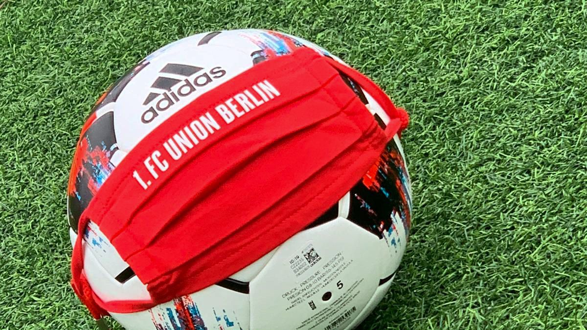 Ein Fußball mit einem roten Mundschutz, auf dem "1. FC UNION BERLIN" steht, liegt auf grünem Kunstrasen.