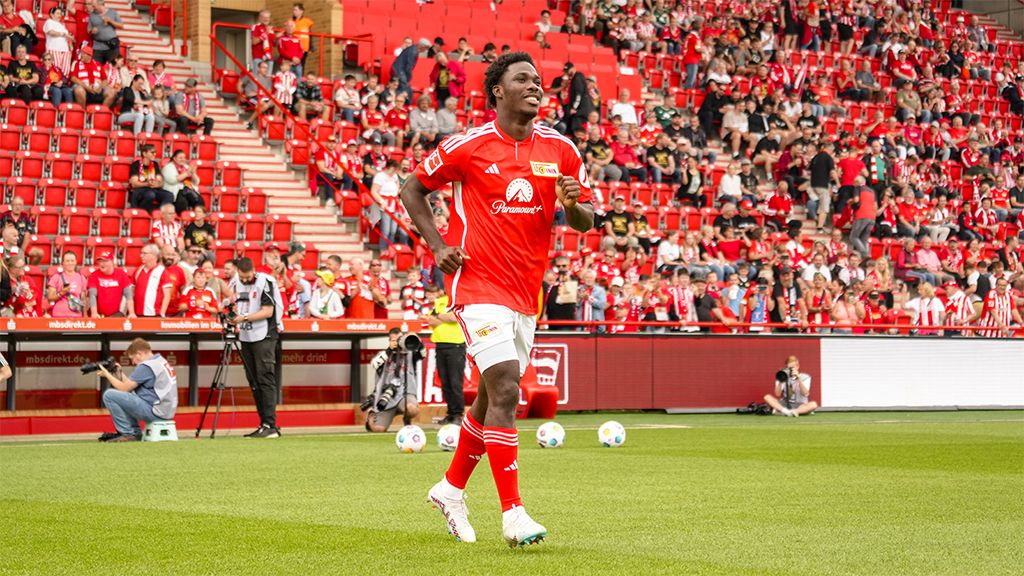 Fußballspieler in rotem Trikot läuft auf dem Spielfeld vor einer begeisterten Menge in einem Stadion.