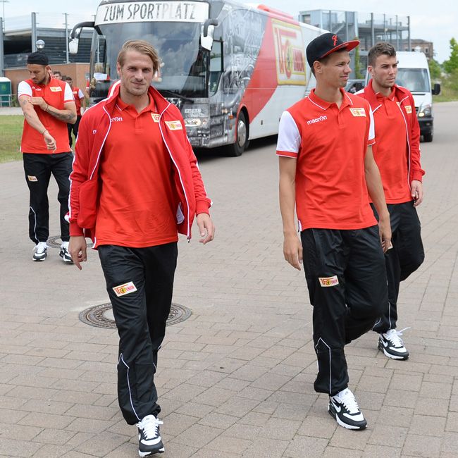 Vier Fußballspieler in roten Trikots und schwarzen Hosen gehen in Richtung eines Busses, umgeben von einem hölzernen Gehweg.