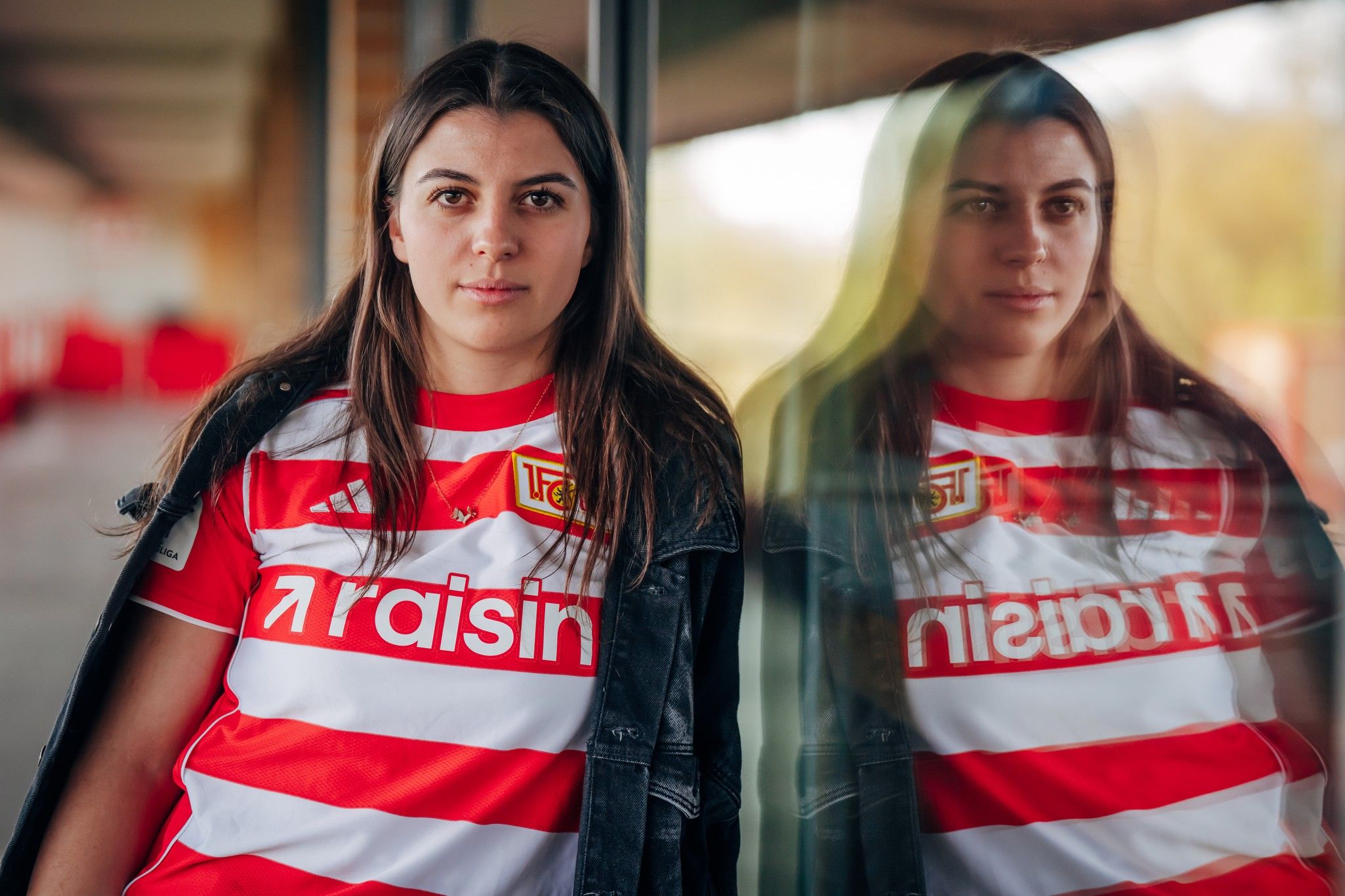 Eine Person mit langen Haaren trägt ein gestreiftes Fußballtrikot und steht vor einer Glaswand, die ihr Spiegelbild reflektiert.