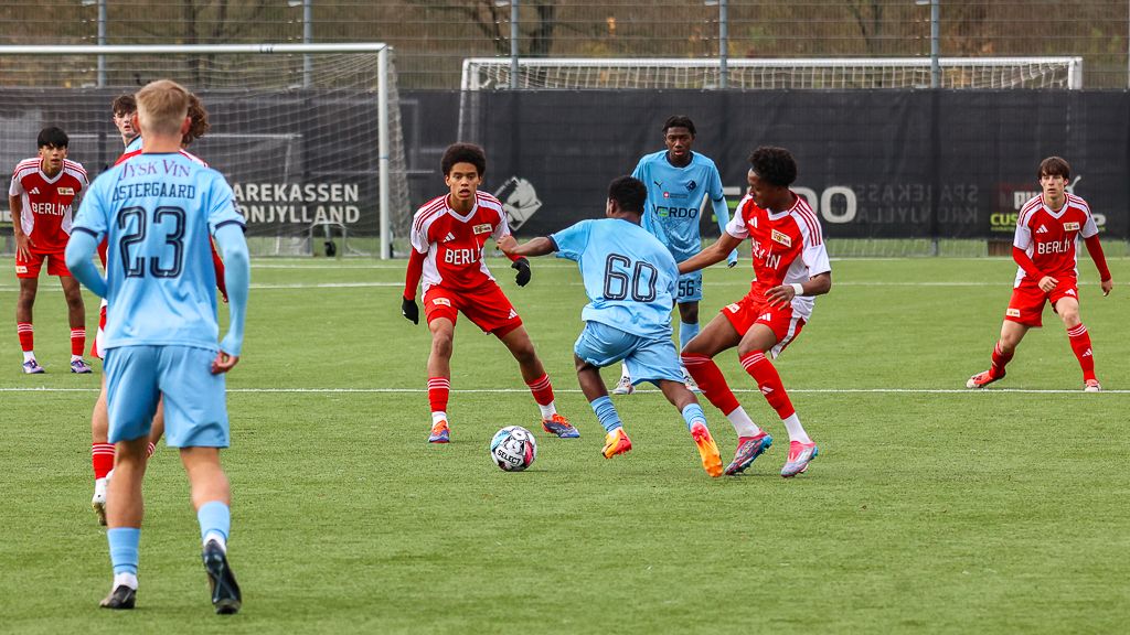 Spielszene eines Fußballmatches zwischen zwei Jugendmannschaften mit Spielern in roten und blauen Trikots auf einem grünen Rasenfeld.