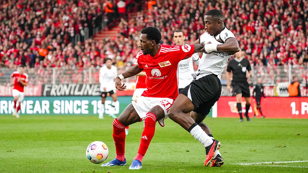 A football player in red fights for the ball against a player in black on the field. Spectators in the background.