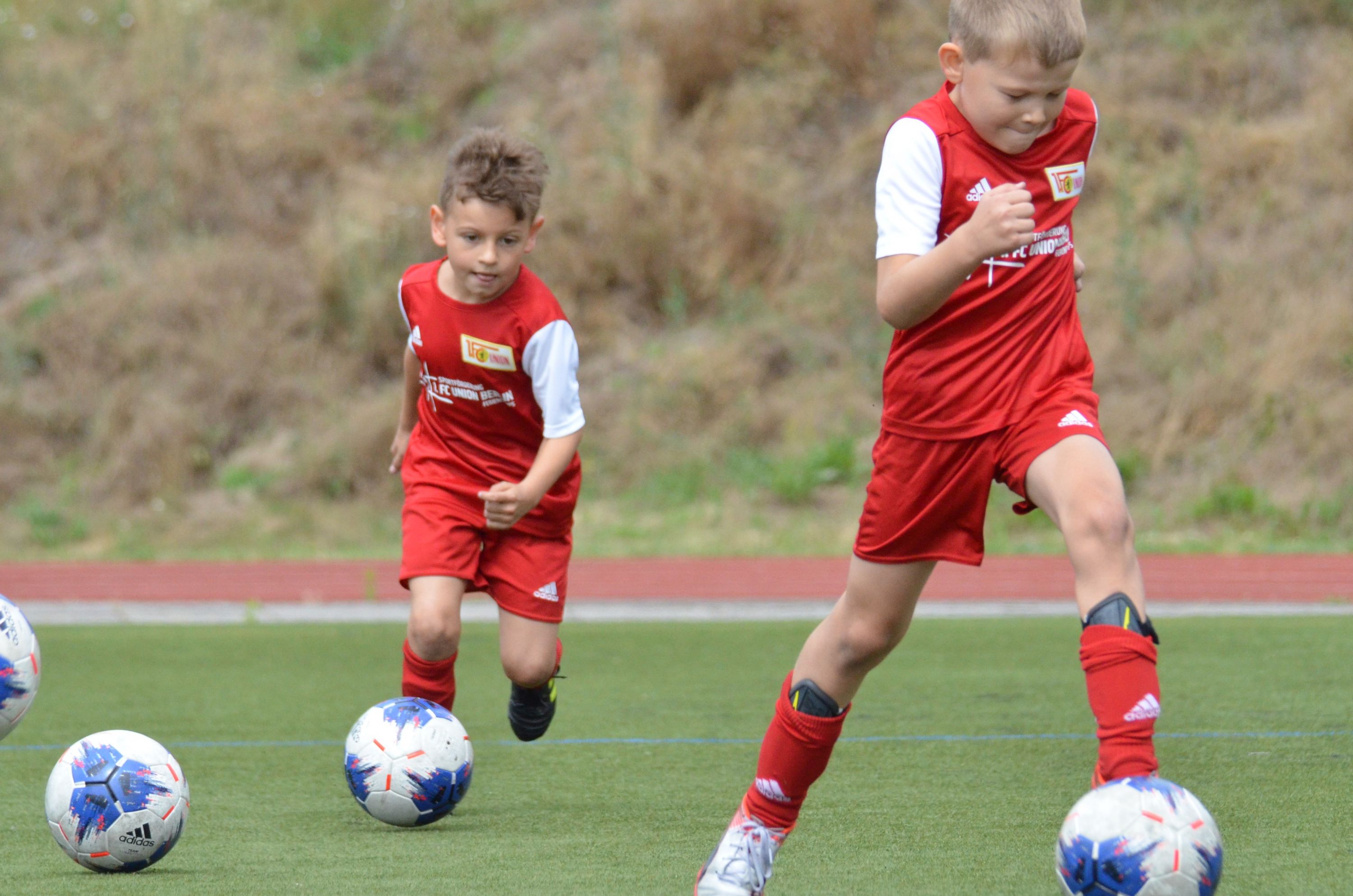 Zwei Jungen in roten Fußballtrikots dribbeln den Ball auf einem Sportplatz.